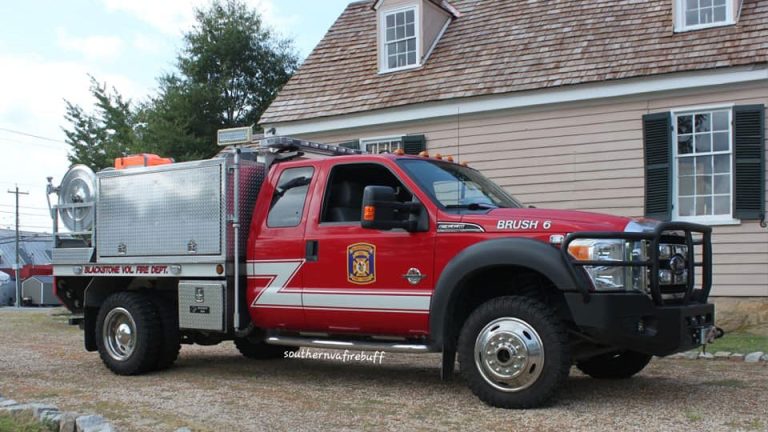 A red fire department truck with "BRUSH 6" on the side is parked in front of a house. The truck has a silver equipment compartment. The words "BLACKSTONE VOL FIRE DEPT" are visible on the side. The house has beige siding and green shutters.