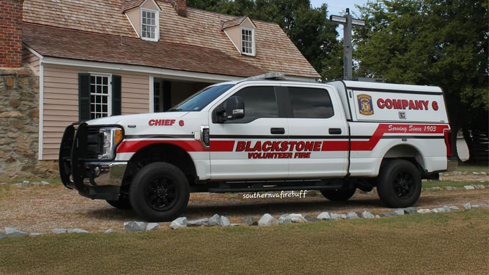 A white Blackstone Volunteer Fire Department truck with red stripes is parked on grass in front of a beige house with a brown shingle roof. The truck is labeled "Chief" and "Company G.