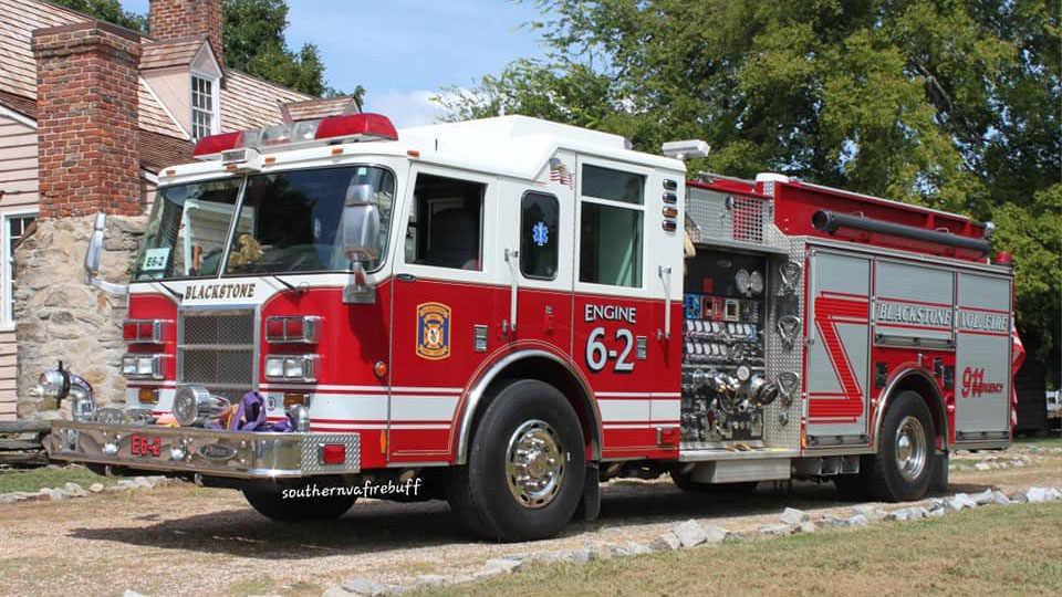 Red and white fire truck labeled "Blackstone Volunteer Fire Department" and "Engine 62" parked in front of a brick house. The truck features multiple hoses and equipment compartments. Trees and a clear sky are in the background.