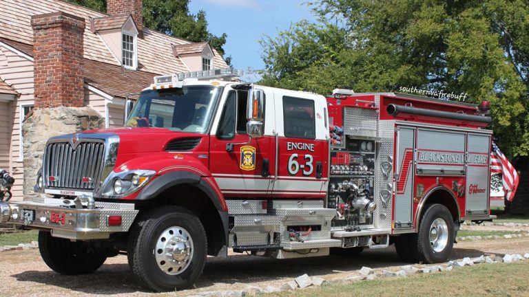 A red and white fire truck labeled "Engine 63" is parked on a driveway near a brick house. The truck has shiny chrome features and visible fire equipment on its side. Trees and a blue sky are in the background.