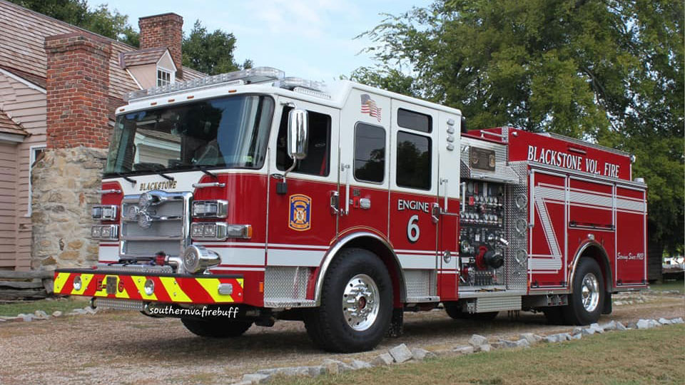 A red and white fire truck labeled "Blackstone Vol. Fire" and "Engine 6" is parked on a grassy area. The truck features various equipment and hoses. A clapboard house with a stone chimney and trees are in the background.