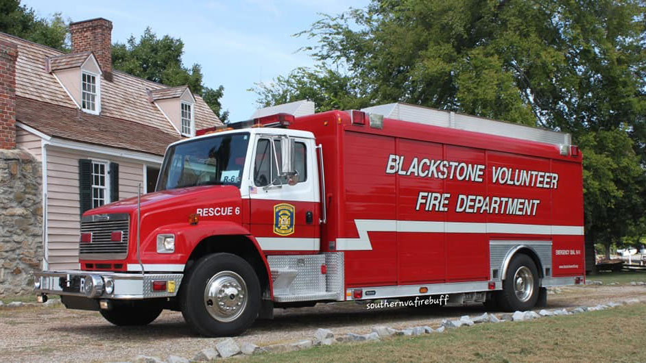 A red fire truck labeled "Blackstone Volunteer Fire Department" is parked on a gravel road beside a historic-style building. The truck has "Rescue 6" marked near the front. Trees are visible in the background.