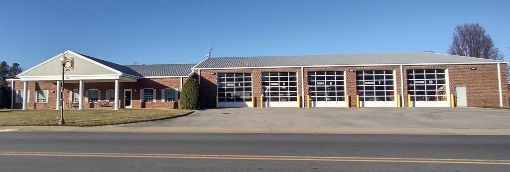 A brick firehouse in Blackstone with a gabled roof features five large garage bays with white doors and yellow bollards. A small lawn and walkway adorn the front, reflecting the community's commitment to volunteer firefighting under a clear blue sky.