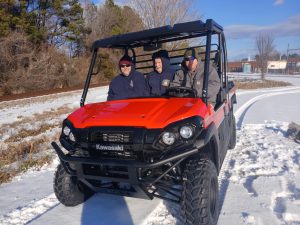 Three people sit in a bright red Kawasaki off-road vehicle on a snowy path. They are dressed in winter clothing. Trees and a building are visible in the background under a clear blue sky.
