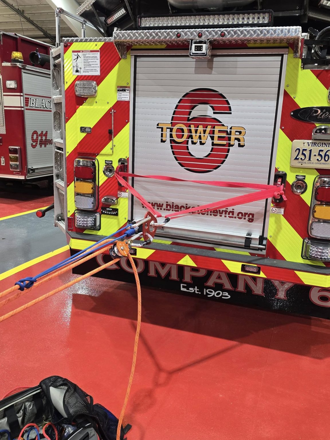 A firetruck with a red and yellow reflective back, labeled "Tower 6", is parked inside a station. Several red straps and pulleys are attached to its rear, extending toward the floor. Equipment bags and tools are visible near the setup.
