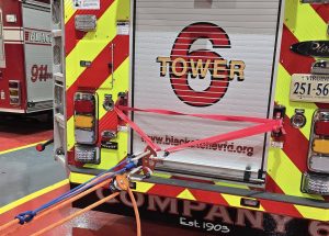 A firetruck with a red and yellow reflective back, labeled "Tower 6", is parked inside a station. Several red straps and pulleys are attached to its rear, extending toward the floor. Equipment bags and tools are visible near the setup.