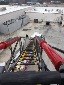 View from an extended fire truck ladder reaching down to an industrial area with a firefighter in a black uniform standing near a fire truck. The ground is concrete with scattered utility equipment. Buildings and a dumpster are visible in the background.
