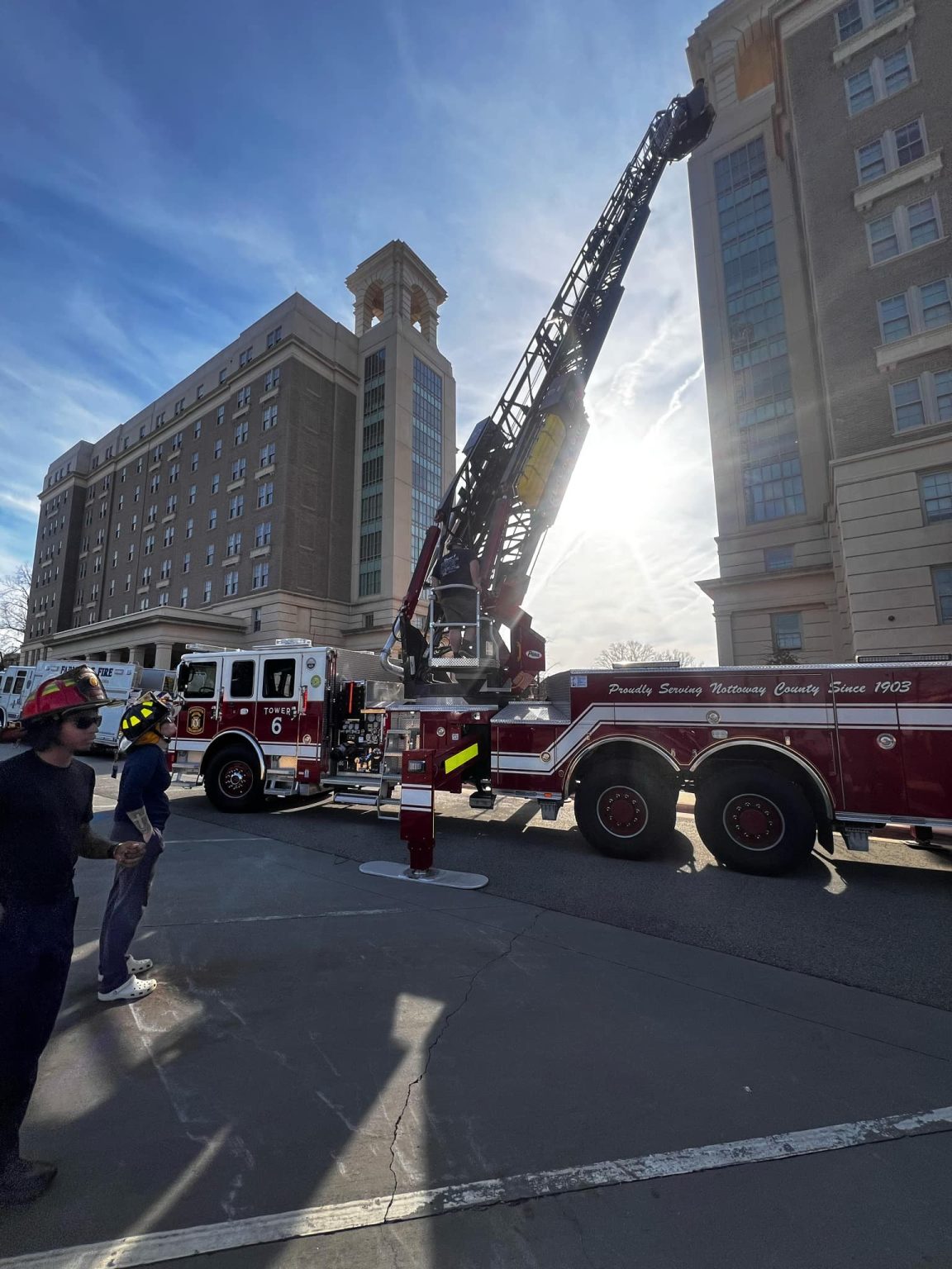 A firetruck with an extended ladder is parked on a city street beside a tall building. The sun is shining brightly in the background. A firefighter stands in the foreground, and there are several other firetrucks nearby.