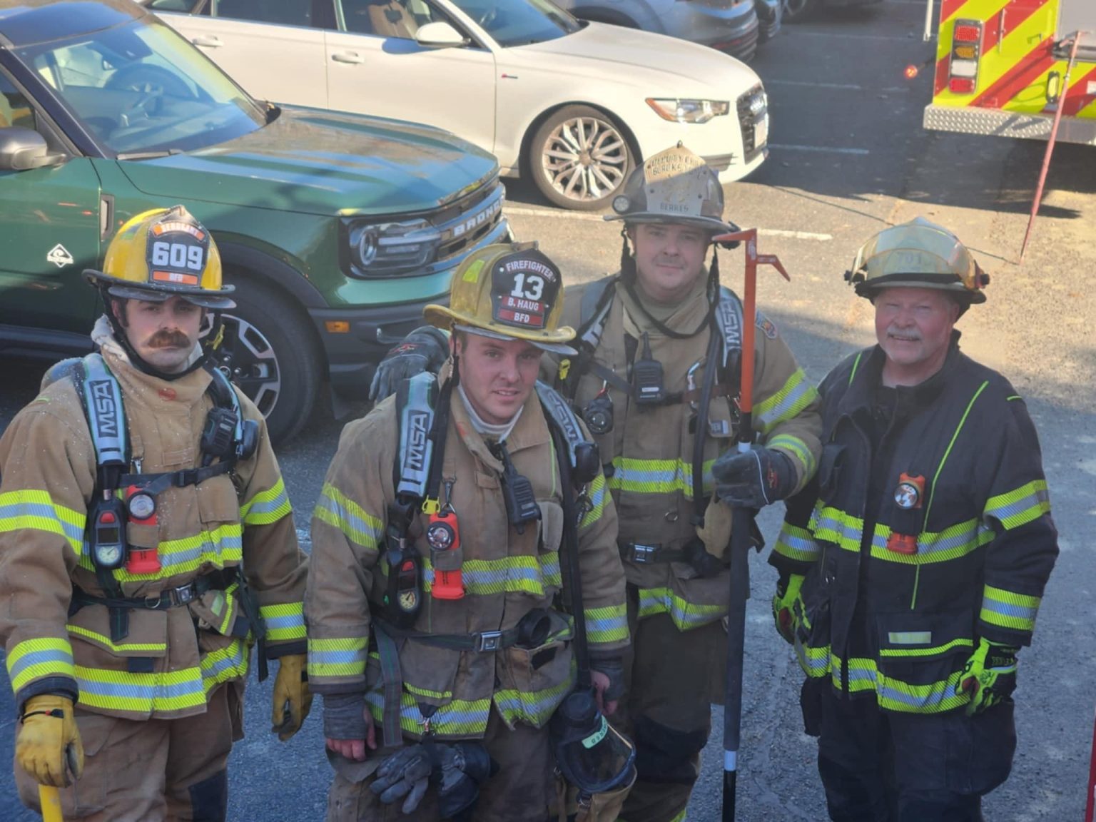 Four firefighters in full gear, including helmets and breathing apparatus, stand together outside. They appear to be on a break, with parked vehicles visible in the background.